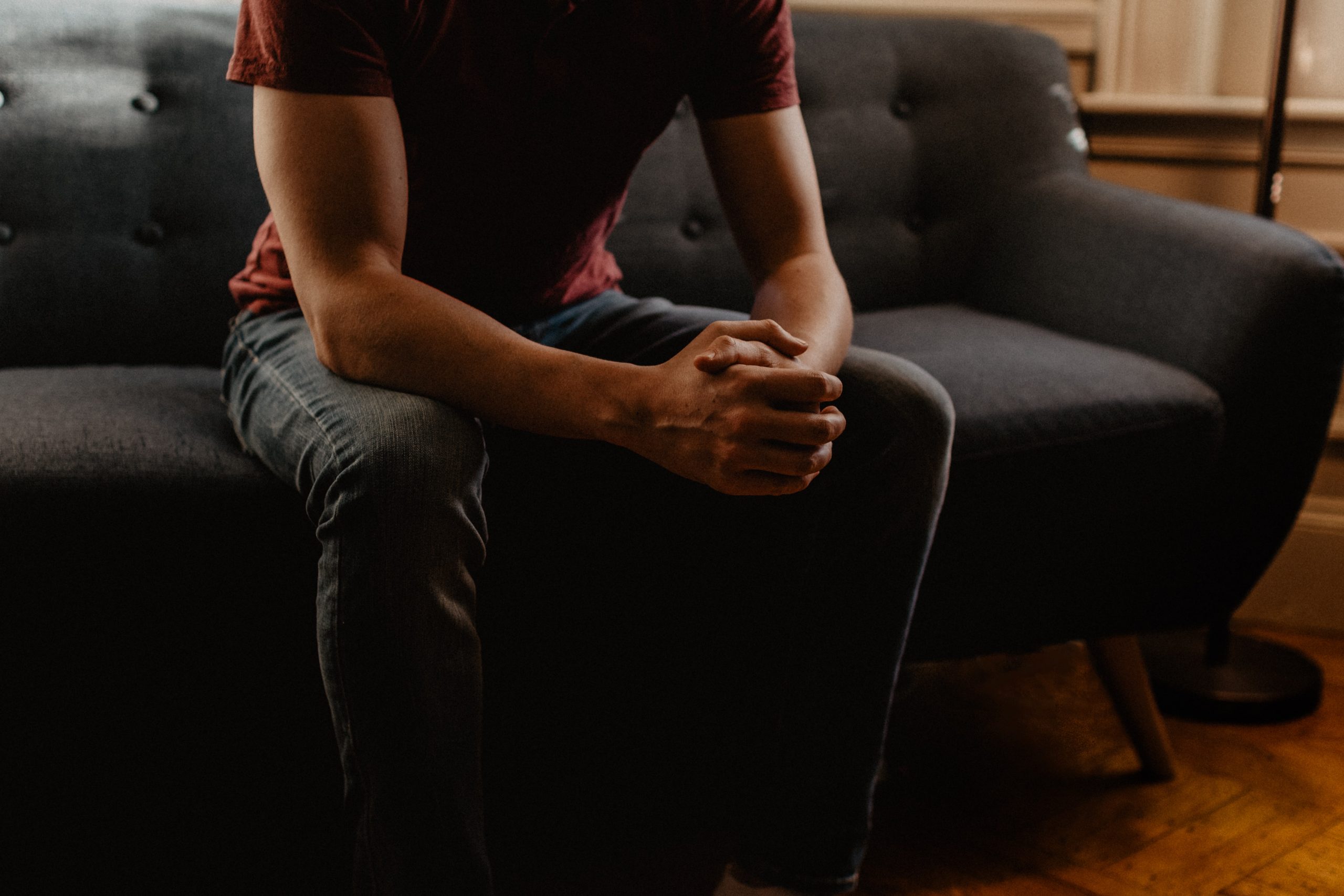 The upper body of a man in a red t-shirt and grey trousers, sitting on a grey couch with his hands clasped together, his arms resting on his thighs