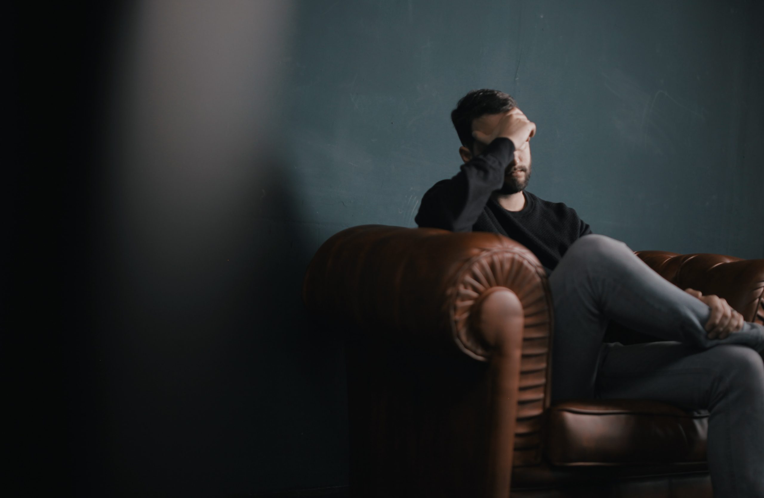 A white man, in a bacl t-shirt and jeans, sitting on a brown, leather couch, resting his hand on his head in contemplation., against a blue/gray wallpaper and an out of focus therapist on the left